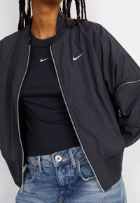 Zwarte bomberjack met rits, ribgebreide manchetten en Nike-logo op de borst, gedragen over een zwart shirt en denim high-waisted shorts.