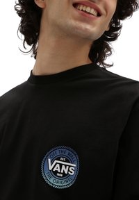Schwarzes T-Shirt mit einem kreisförmigen Vans-Logo in Blau und Weiß, mit dem Text "Off the Wall" und "Established 1966." Aus weichem Baumwollmaterial.