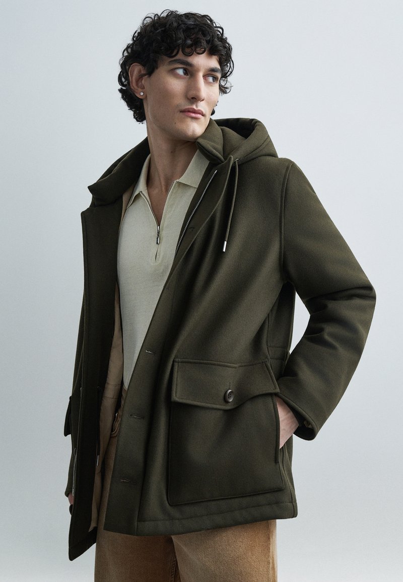 Oliivinvihreä hupullinen takki, jossa on pehmeä pinta, etutaskut ja napillinen sulkus, kerroksittain päälle puetun vaalean beigen vetoketjullisen paidan kanssa.