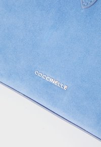 Kék velúr kézitáska sima textúrával, ezüst színű, dombornyomott "Coccinelle" márkanevével és minimalista kivitelben.