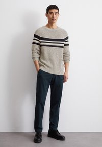 Beige gestrickter Pullover mit schwarz-weißen Streifen, kombiniert mit dunkelgrünen Hosen und schwarzen Schuhen. Glatte Textur, lockere Passform.