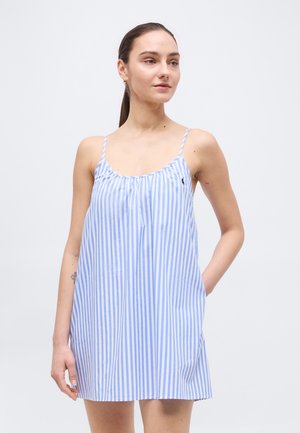 Žena nosi modré a biele šaty s tenkými ramienkami a vertikálnym pruhovaním, stojaca s jednou rukou v vrecku na jednoduchom pozadí.