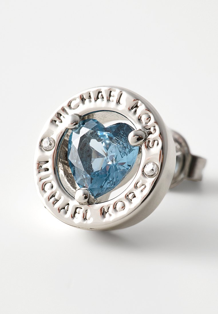 Niebieski kamień szlachetny w kształcie serca osadzony w okrągłej srebrnej obręczy z wygrawerowanym napisem "Michael Kors" oraz dwoma przezroczystymi akcentami po bokach.