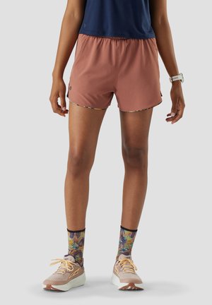 Persoon met roestkleurige shorts, meerkleurige bloemenprint sokken, beige hardloopschoenen, marineblauw shirt en een wit horloge, staand op een grijze achtergrond.