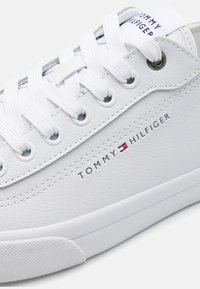 Sneaker sintetico bianco con superficie testurizzata, dotato di lacci bianchi piatti, logo "TOMMY HILFIGER" stampato in nero e suola in gomma.