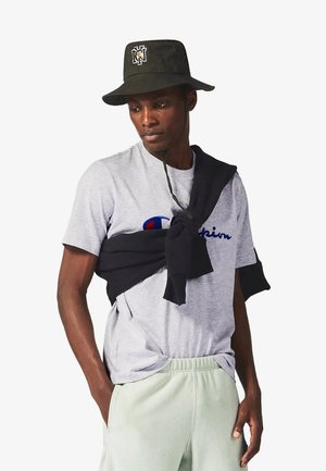 Uomo con cappello a secchiello nero, maglietta grigia Champion, pantaloni chiari e felpa nera legata sulle spalle, che guarda verso il basso e di lato.