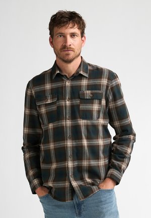 Mand med kort brunt hår og skæg iført en mørkegrøn og brun ternet skjorte med knapper med hænderne i lommerne på lyseblå jeans.