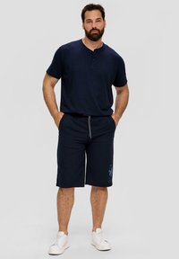 Marineblauwe casual overhemd met henley-halslijn, gecombineerd met bijpassende shorts met een trekkoord en bloemenprint. Witte sneakers maken de look af.