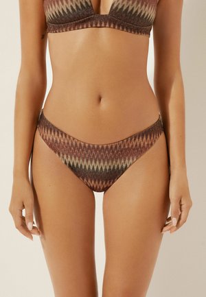 Kvinde iført brun bikini med zigzag-mønster, der viser torso og øvre lår mod en ensfarvet baggrund.