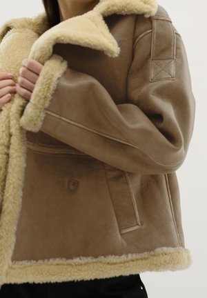 Person iført en brun shearling-jakke med cremefarvet fleecefor, holder i kraven og viser detaljeret syning på ærmer og lommer.