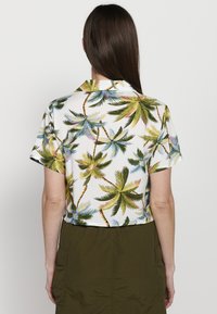 Gekürztes Shirt aus weißem Stoff mit einem Muster von grünen und blauen Palmen. Kurze Ärmel und ein klassischer Kragen.