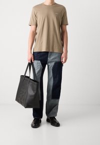 Mand iført en beige Hugo T-shirt, patchwork blå denim bukser, sorte sko, der holder en stor sort læder tote taske med præget logo.