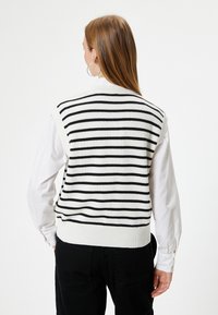 Chaleco de punto a rayas en blanco y negro con dobladillo acanalado, usado sobre una camiseta de manga larga blanca, presenta un diseño sin mangas.