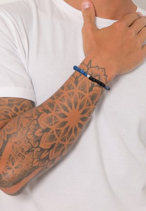 Mønstret armbånd med blå, sorte og hvide perler, båret på et tatoveret håndled over en hvid t-shirt. Glatte, runde perler.