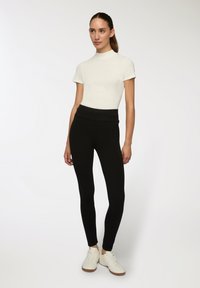 Biała bluzka z krótkim rękawem w połączeniu z czarnymi legginsami o wysokim stanie, charakteryzującymi się gładką teksturą. Model nosi białe sneakersy z brązową podeszwą.