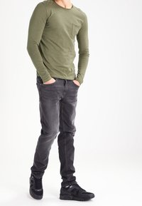 Camisa de manga larga verde oliva con bolsillo en el pecho, combinada con vaqueros gris oscuro y zapatillas negras con detalles en blanco.