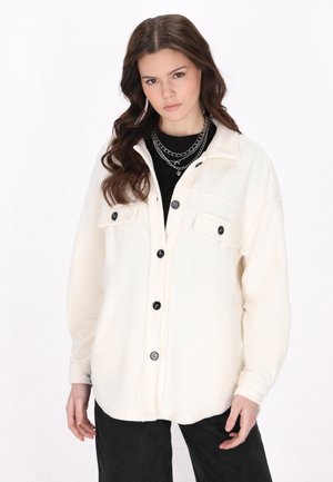 Hvid fleece skjortejakke med to brystlommer, sorte knapper og krave, parret med en sort top og mørke bukser.