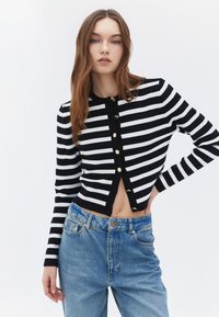 Randig kavaj i svart och vitt med en kort design, rund halsringning och guldknappar, kombinerad med högmidjade blå jeans.