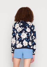 Person med lockigt hår som bär en marinblå blommig långärmad topp och ljusblå shorts, stående mot en enfärgad vit bakgrund.