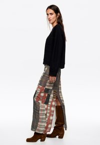 Suéter negro de corte relajado, combinado con una larga falda a cuadros en gris, rojo y beige, que presenta aberturas laterales y botas de tacón marrones.