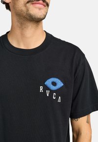 Camiseta de algodón negra con un diseño de ojo bordado en azul y texto estilizado en blanco en el lado izquierdo del pecho. Tiene mangas cortas y un cuello redondo.