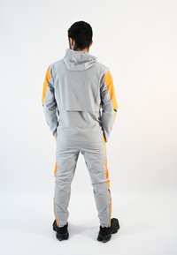 Chándal deportivo gris con detalles en naranja. Presenta una capucha, bolsillos con cremallera y un diseño ajustado. Se lleva con zapatillas deportivas negras.