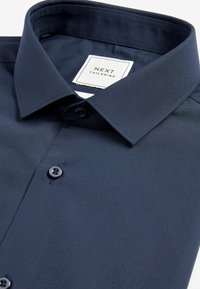 Marineblå skjorte med knapper, standard krave, glat tekstur, to sorte knapper og en mærkat med teksten "NEXT TAILORING."