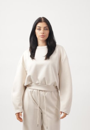 Kvinde med langt mørkt hår iført en løs beige sweatshirt og matchende bukser med snøre, stående foran en ensfarvet hvid baggrund.