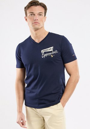 Jeune homme debout, une main dans la poche, portant un T-shirt bleu marine à col en V avec le logo "Armorlux Equipage & Co." et un pantalon beige.