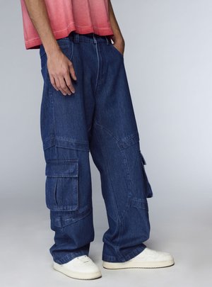 Persona che indossa pantaloni cargo in denim blu scuro larghi con grandi tasche, sneakers bianche e una maglietta corta rossa, in piedi con le mani nelle tasche.