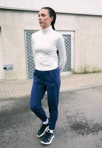 Biała, zapinana na zamek kurtka sportowa, niebieskie dopasowane dresy, czarne sneakersy z białą podeszwą; odzież z markowym logo Nike.