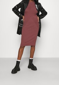 Ribbad, figurnära midi-klänning i djup lila med ett strukturerat mönster. Kombinerad med en skinande svart läderjacka och kraftiga svarta snörstövlar.