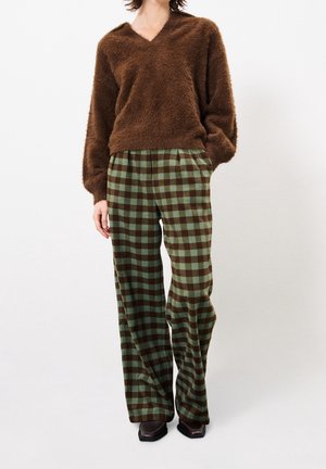 Osoba oblečená v hnědém chlupatém svetru s výstřihem do V a zelenohnědých kostkovaných kalhotách s širokými nohavicemi a černých botách, stojící před bílým pozadím.