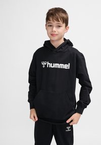 Schwarzer Hoodie aus weichem Stoff mit einer Fronttasche, der ein weißes "hummel"-Logo auf der Brust trägt, sowie passenden schwarzen Hosen.