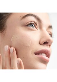 Fondotinta in una tonalità beige cremosa applicato su pelle chiara. Texture liscia, primo piano di una mano che stende delicatamente il prodotto sulla guancia.