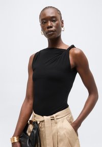 Γυναίκα φοράει μαύρο αμάνικο τοπ, μπεζ ψηλόμεσο παντελόνι με ζώνη, χρυσά σκουλαρίκια κρίκους, βραχιόλι και κρατά μια μαύρη τσάντα.