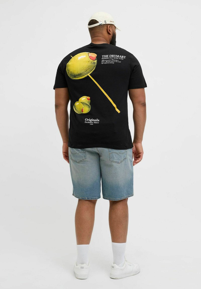 Schwarzes T-Shirt mit kurzen Ärmeln, auf dem die gelben Oliven und ein Spieß-Design auf der Rückseite abgebildet sind. Kombiniert mit hellblauen Denim-Shorts und weißen Sneakers.