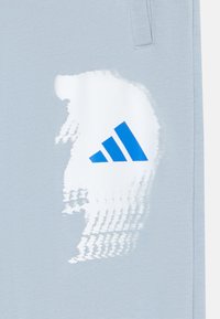 Világoskék sportnadrág, fehér textúrázott grafikával és kék háromcsíkkal ellátott logóval a bal oldalon. Puha anyag, laza szabás.