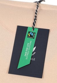 Lichtbeige katoenen kledingstuk met een gestructureerd oppervlak en een groene Fair Trade-label bevestigd met een zwart-witte draad.