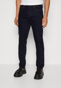 Vaqueros de mezclilla azul oscuro con costuras en naranja contrastante, diseño de corte ajustado, estilo estándar de cinco bolsillos, y combinados con unos mocasines negros gruesos.