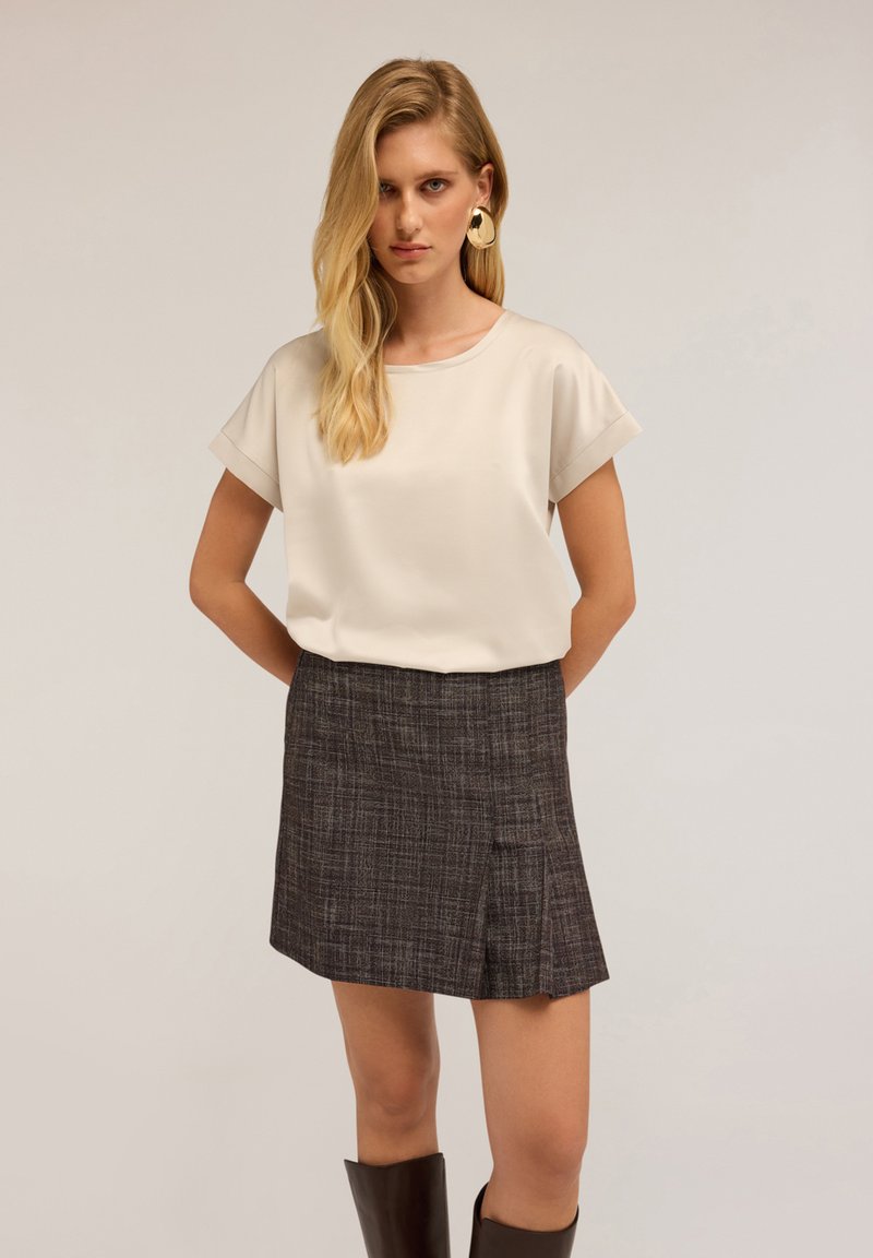 Blusa beige a maniche corte abbinata a una minigonna tartan scura con una piega laterale. Modella con orecchini rotondi audaci e stivali al ginocchio marroni.