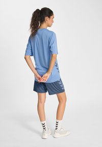 Vrouw met krullend haar in een paardenstaart, gekleed in een lichtblauwe sportshirt en donkerblauwe shorts, staande met de handen achter haar rug gekruist.