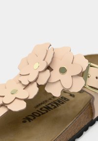 Κοντινή λήψη του μπεζ λουριού σανδαλιού Birkenstock με στρωματοποιημένες, λουλουδένιες δερμάτινες λεπτομέρειες και χρυσούς καρφίτσες.