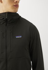 Črna jakna s polnim zapiranjem in visokim ovratnikom, teksturirana tkanina ter izstopajoč logotip Patagonia na prsih. Ima vrvice za zategovanje in zadrgo za zapiranje.