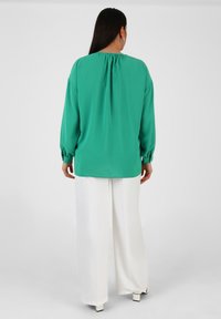 Grüne Bluse mit gefaltetem Rückendesign, langen Ärmeln mit elastischen Bündchen, kombiniert mit weißen weitgeschnittenen Hosen. Glattes, leichtes Material.