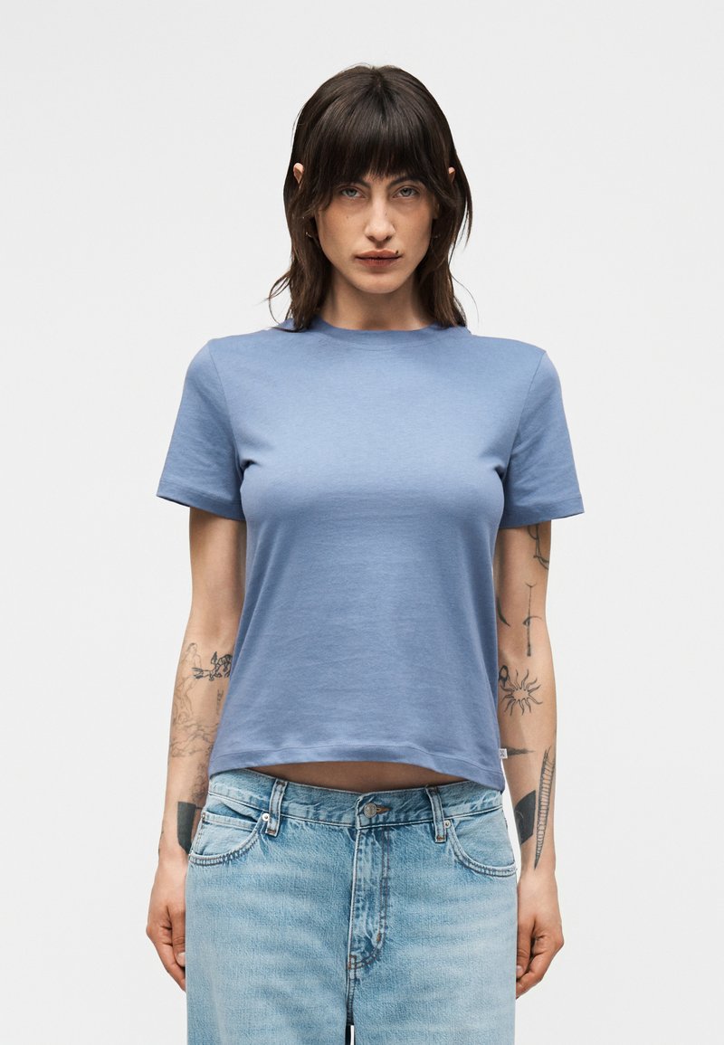 Γυναίκα με σκούρα μαλλιά μήκους μέχρι τους ώμους φοράει ένα απλό ανοιχτό μπλε κοντομάνικο t-shirt και ανοιχτό μπλε τζιν, στέκεται μπροστά σε ένα απλό φόντο.