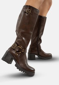 Botas de cuero marrón hasta la rodilla con un tacón ancho, decoradas con hebillas y correas, que presentan un cierre de cremallera y una suela texturizada para mayor agarre.