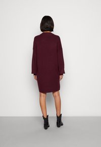 Burgunderfarbendes Strickkleid mit langen Ärmeln, lockerer Passform und rundem Ausschnitt, kombiniert mit schwarzen Knöchelstiefeln. Glatte Textur; knielanges Design.