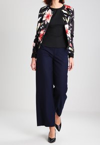 Veste bomber noire à motif floral avec des accents roses et verts, associée à un haut noir ajusté et un pantalon large bleu marine. Talons pointus noirs.
