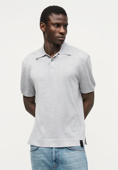 Άνδρας που φορά ανοιχτό γκρι κοντομάνικο πόλο μπλουζάκι και μπλε τζιν, στέκεται με τα χέρια πίσω από την πλάτη, κοιτάζοντας προς τα αριστερά του.